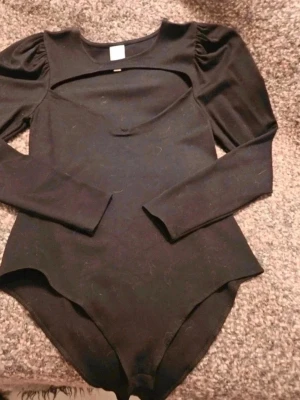 Svart body med cut out H&M L - Säljer en svart body med lång ärm och cool cut out-detalj över bröstet. Modellen har puffiga axlar. Tyget är mjukt och stretchigt, perfekt till en edgy outfit. Då jag fotograferade bodyn på en alpacka filt så blev den hårig men kommer så klart rollas och skickas i ny skick  Kan även hämtas I Partille  E5