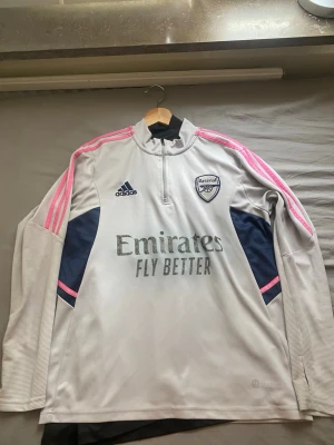 Arsenal träningströja  - Snygg långärmad träningströja från Arsenal i ljusgrått med mörkblå detaljer och rosa ränder på ärmarna. Halv dragkedja framtill, . Perfekt för fotbollsträning eller gym, som ni ser på bilderna så har texten på tröjan gått bort men inget som märks 