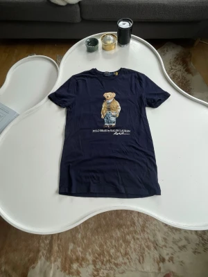 Mörkblå Polo Bear t-shirt Ralph Lauren - Säljer en mörkblå t-shirt från Ralph Lauren med den ikoniska Polo Bear-trycket på bröstet. T-shirten har rund hals och korta ärmar, tillverkad i mjuk bomull för skön känsla. Perfekt för dig som gillar klassiska streetwear-vibbar. Custom slim-fit