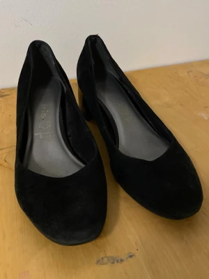 Svarta mockapumps med låg klack - Ett par klassiska svarta pumps i mocka med rundad tå och låg, stabil klack. Skorna har en stilren design och är tillverkade i ett mjukt material som ger en elegant känsla. Perfekta för dig som gillar en tidlös och enkel look.