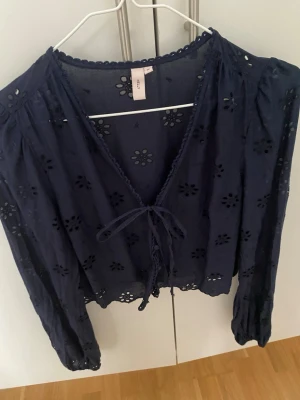 Mörkblå blus med broderade blommor - Söt mörkblå blus med broderade blommor och hålmönster. Blusen har v-ringning med knytband framtill, vågig kant och långa ärmar med resår vid handlederna. Perfekt för en trendig och avslappnad look.