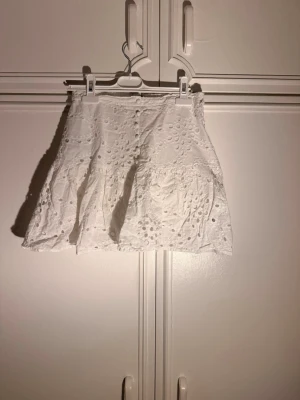 Vit broderad minikjol med volang - En vit kjol med ”hål” och underkjol för säkerhetsskull, det finns även fyra knappar på den. Alla detaljer på kjolen gör den lite extra fin och det är det som lockade mitt intresse. Finns även en dragkedja på sidan av kjolen! Den har använts några gånger på sommaren men inte så mycket efter det🤍