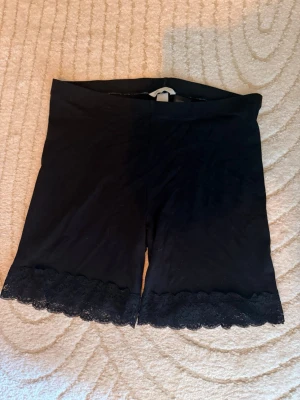 Svarta shapewear-shorts med spets - Säljer ett par svarta shapewear-shorts med bred resår i midjan och fina spetsdetaljer längst ner på benen. Shortsen har en mjuk och stretchig känsla som formar och ger stöd. Perfekta under klänning eller kjol för en slät siluett.