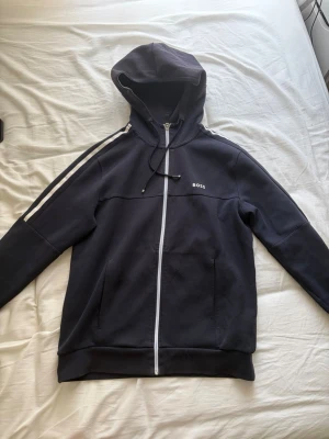 Svart hoodie med dragkedja från BOSS - Snygg svart hoodie från BOSS med vit dragkedja och vita ränder längs ärmarna. Klassisk huva med snörning och två fickor framtill. Perfekt för en chill och sportig look.