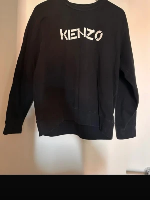 Svart sweatshirt från Kenzo - Säljer en svart sweatshirt från Kenzo med logga i vitt framtill. Tröjan har rund halsringning och långa ärmar. Perfekt för dig som gillar streetwear och vill ha en enkel men snygg look.