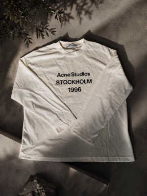 Vit långärmad topp Acne Studios - Vit långärmad topp från Acne Studios med svart text 'Acne Studios STOCKHOLM 1996' på bröstet. Tillverkad i mjuk bomull med klassisk passform och rund halsringning. Perfekt för en clean och stilren look.