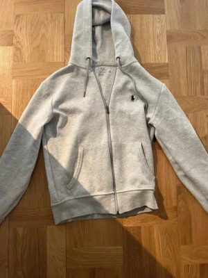 Grå zip hoodie från Polo Ralph Lauren - Snygg grå hoodie med dragkedja från Polo Ralph Lauren. Klassisk design med huva, fickor framtill och den ikoniska lilla loggan broderad på bröstet. Tillverkad i mjukt material som känns skönt mot huden. Perfekt för en avslappnad och stilren look.