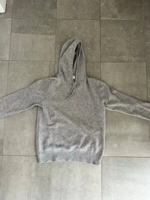Grå cashmire  hoodie  - En stilren grå hoodie med huva och dragsko. Tröjan har långa ärmar och ribbade muddar vid ärmslut och nederkant. Perfekt för en avslappnad look och passar till det mesta. Materialet känns mjukt och bekvämt mot huden. Bra skick dock ett litet hål på ena ärmen vid armbågen men de går att fixa. Om du undrar något är de bara att höra av sig!