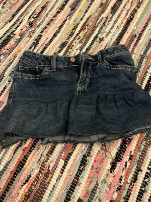 Mörkblå jeanskjol med volang - Säljer en egensydd mörkblå jeanskjol med volangkant nedtill och markerade vita sömmar. Kjolen har klassiska jeansfickor fram och bak samt bälteshällor och knappstängning. Perfekt för dig som gillar en cool och avslappnad stil. Tyget är från jeans från Lindex!