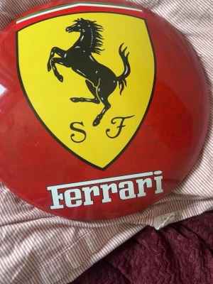 Rund Ferrari-väggskylt i metall - Säljer en rund väggskylt i metall med klassiska Ferrari-loggan i gult, svart och rött. Skylten har den svarta hästen och texten 'Ferrari' i vitt. Perfekt för dig som gillar bilar och vill ha något snyggt på väggen.