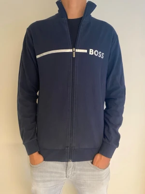 Mörkblå ziptröja från BOSS - Snygg mörkblå ziptröja från BOSS med vit logga och rand över bröstet. Tröjan har hög krage, hel dragkedja och ribbade muddar vid ärmslut och nederkant. Perfekt för en stilren och avslappnad look.