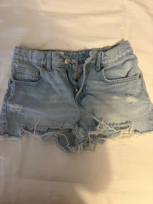 Jeans shorts från Zara kids - Säljer ett par ljusblå jeansshorts i storlek 13-14  med medel hög  midja och råa, fransiga kanter. Shortsen har slitningar framtill och klassiska fem fickor. Perfekta för varma dagar och en avslappnad stil. 