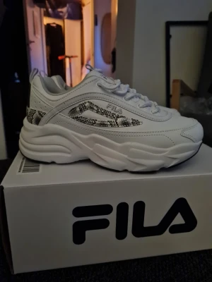 Vita chunky sneakers från FILA, NYA - Helt nya snygga vita sneakers från FILA, stl 37, med chunky sula och detaljer i ormskinnsmönster. Skorna har en modern siluett, vita skosnören och är tillverkade i syntetmaterial med coola kontraster. Perfekta för dig som vill sticka ut med din stil.