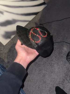 Svart Gucci keps med ormtryck - Säljer en svart keps från Gucci med klassiskt GG-mönster och en stor röd orm på framsidan. Kepsen har mesh på sidorna och justerbar rem bak. Materialet är en mix av canvas och mesh vilket ger en cool streetwear-look.