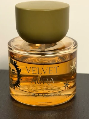 Velvet Aura parfym från Rituals - Velvet Aura från Rituals är en exklusiv parfym i en rund glasflaska med sol- och stjärndetaljer. Flaskan har en olivgrön, matt kork och innehållet är bärnstensfärgat. Designen känns lyxig och unik, perfekt för dig som gillar snygga detaljer på parfymflaskan. Den är lite använd som ni ser. Skulle säga att ca 5-10 ml av flaskan är använd