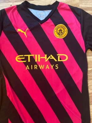 Manchester City matchtröja  - Snygg Manchester City matchtröja från Puma i svart med rosa diagonala ränder och gula detaljer. Tröjan har klubbmärke och sponsortryck på bröstet samt korta ärmar. Tillverkad i ett lätt och funktionellt material som passar perfekt till träning eller match.