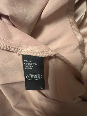 Ljusrosa klänning från Cider - Säljer en ljusrosa klänning från Cider i storlek L. Klänningen har lång ärm, v-ringning och krage. Materialet känns lent och stretchig och har ett lätt glansigt utseende, vilket ger en elegant touch. Kjoldelen är veckad för extra volym.