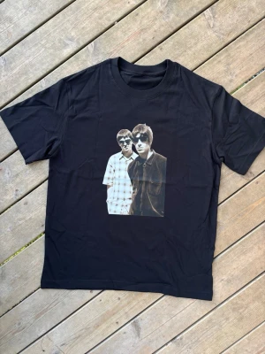 Oasis Noel & Liam merch vintage Svart t-shirt med fototryck - Svart oasis tshirt i storlek s. 100% bomull bra kvalitet. Unisex. Längd ca 69cm axelbredd ca 47,5cm. Ny. Skickar gärna i bundle. Cool svart t-shirt med ett stort fototryck av två personer med solglasögon på bröstet. T-shirten har klassisk passform, rund hals och är tillverkad i mjukt bomullsmaterial. Perfekt för dig som gillar grafiska motiv och streetwear.