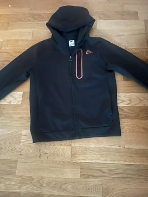 Nike tech fleece hoodie - svart/Orange - xl (158-170cm) - Säljer en Nike tech fleece hoodie i fint skick. Alla dragkedjor fungerar och den har inga större skador. Svart med orange detaljer. (Storlek XL 158-170 cm) hör gärna av dig om du har frågor eller vill ha fler 