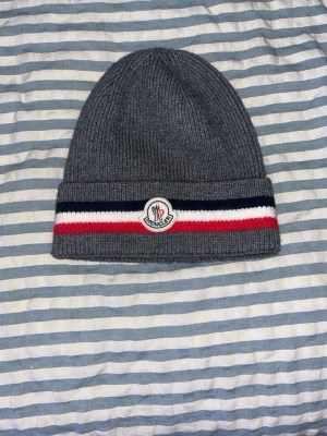 Grå Moncler mössa med ränder - Snygg grå ribbstickad mössa från Moncler med vikbar kant. Mössan har breda ränder i mörkblått, vitt och rött samt Moncler-logga framtill. Perfekt för dig som vill ha en klassisk men ändå sportig look. Tillverkad i mjukt stickat material.