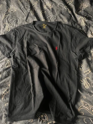 Svart t-shirt från Polo Ralph Lauren - Klassisk svart t-shirt från Polo Ralph Lauren med röd broderad logga på bröstet. T-shirten har rund halsringning och korta ärmar. Tillverkad i mjuk bomull som känns skön mot huden. Perfekt basplagg för alla tillfällen.