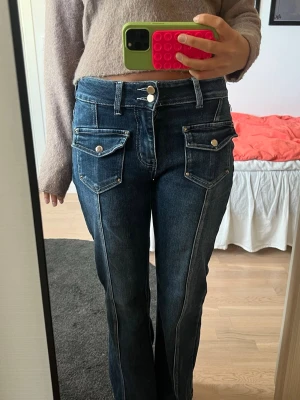 Mörkblå jeans med coola fickor - Mörkblå raka jeans med coola fickor och vita sömmar. Fickor med lock och silverknappar. Jeansen är låga i midjan. Materialet är bomull, polyester och elastan.