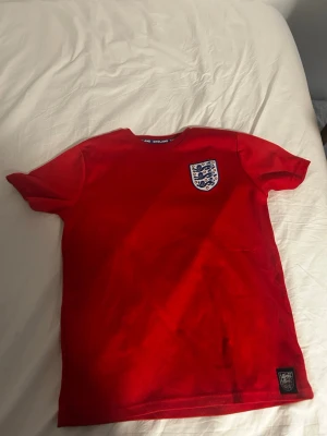 Röd England fotbollströja - Snygg röd fotbollströja med England-emblem på bröstet. Tröjan har korta ärmar och rund hals. Tillverkad i ett lätt och ventilerande material som passar perfekt till träning eller match. Klassisk design med broderad logga framtill.