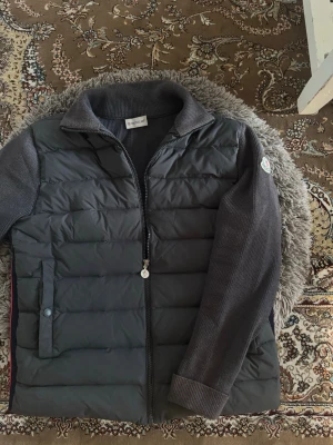 Moncler cardigan - Snygg svart dunväst från Moncler med stickade mörkgrå ärmar och krage. Västen har dragkedja framtill, två fickor med knapp och en Moncler-logga på ärmen. Perfekt för lager-på-lager och streetstyle.