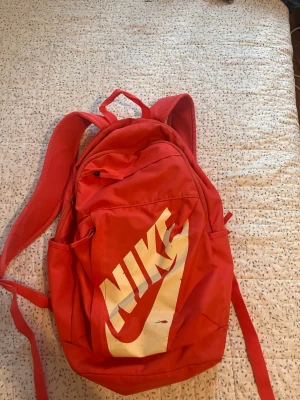 Röd ryggsäck från Nike - Säljer en röd ryggsäck från Nike med stort vitt logotryck på framsidan. Ryggsäcken har justerbara axelband, bärhandtag och flera fack med dragkedja. Tillverkad i slitstarkt syntetmaterial, perfekt för skolan eller träningen.