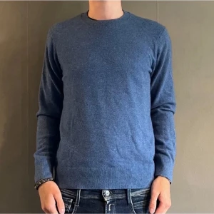 Blå rundhalsad stickad tröja - En stilren blå stickad tröja med rund halsringning och långa ärmar. Tröjan har ribbade muddar vid ärmslut och nederkant, vilket ger en klassisk och enkel look. Perfekt att matcha med jeans för en avslappnad stil.