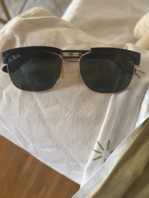 Svarta Clubmaster solglasögon Ray-Ban - Snygga svarta Clubmaster solglasögon från Ray-Ban med klassisk halvbåge och gulddetaljer. Glasen är mörka och bågarna har en retroinspirerad form som passar till alla stilar. Perfekta för soliga dagar och ger en cool vibe. Gamla men i fint skick inga tydliga repor . Kommer utan box 