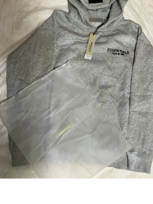 Grå Essentials Fear of God hoodie - Snygg grå hoodie från Essentials Fear of God med svart logga tryckt på både bröstet och ryggen. Klassisk huva och känguruficka framtill. Tillverkad i mjukt bomullsmaterial som känns skönt mot huden. Perfekt för en avslappnad streetwear-look.