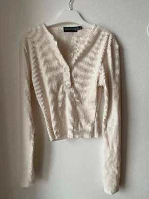 Ribbad beige långärmad topp - Säljer en ribbad beige långärmad topp från PrettyLittleThing. Modellen har knappar framtill och en lätt croppad passform. Perfekt till jeans eller kjol för en enkel men snygg look. Materialet är mjukt och stretchigt för extra komfort.