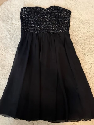 Svart axelbandslös klänning från Vila - Svart axelbandslös klänning från Vila med glittrig paljettöverdel och lätt, flödande kjol. Perfekt för dig som vill sticka ut med en elegant look. Klänningen har en klassisk passform och är knälång.