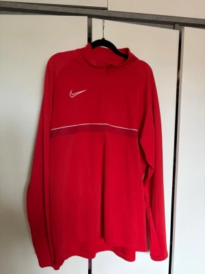 Röd Nike Dri-Fit träningströja - Röd långärmad träningströja från Nike med halv dragkedja och vit logga på bröstet. Tillverkad i Dri-Fit material som transporterar bort fukt. Tröjan har en vit linje över bröstet och är perfekt för sport eller träning.