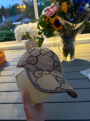 Beige keps  - Säljer en beige keps från ett monogramt i mörkbrunt över hela kepsen. Kepsen har böjd skärm och bruna kanter. Tillverkad i ett slitstarkt tyg som känns lyxigt och trendigt. Perfekt för dig som vill ha en statement-accessoar.
