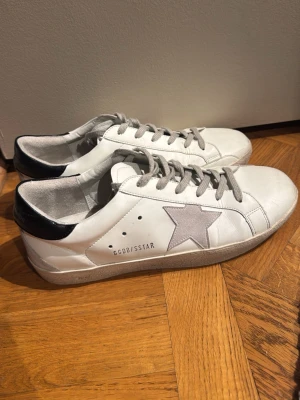 Golden goose skor  - Snygga vita sneakers från golden goose med grå stjärna på sidan och grå skosnören. Skorna har svart hälparti och perforerade detaljer vid sidan. Klassisk låg modell med rund tå och platt sula, tillverkade i läder. Tveka inte på att höra av er vid frågor! Köpta på vongreven closet!