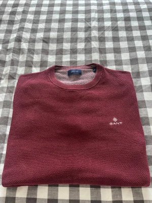 Vinröd tröja från GANT - Snygg vinröd tröja från GANT med rund halsringning och diskret logga broderad på bröstet. Tröjan har en strukturerad yta och ribbade muddar. Perfekt för dig som gillar klassisk och stilren design.
