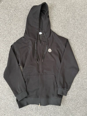 Svart hoodie med dragkedja - Snygg svart hoodie med dragkedja framtill och klassisk M-logga på bröstet. Hoodien har huva med dragsko, ribbade muddar och två stora fickor framtill. Tillverkad i mjuk bomull för skön känsla och stilren look.