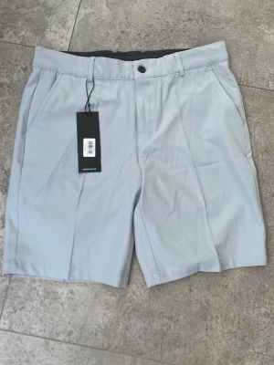 Ljusgrå shorts från Druids - Helt nya Snygga ljusgrå shorts från Druids med klassisk passform och knappstängning i midjan. Shortsen har sidofickor och bälteshällor, perfekta för en stilren och enkel look. Tillverkade i ett mjukt och lätt material som passar bra till varmare dagar.