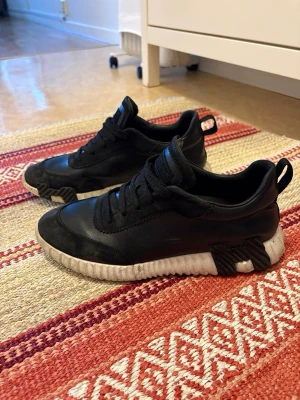Svarta sneakers med vit sula - Säljer ett par svarta sneakers med chunky vit sula och svarta detaljer. Skorna har en låg profil, snörning och dragflik bak. Materialet ser ut att vara en mix av läder och syntet. Perfekta för en streetwear-look.