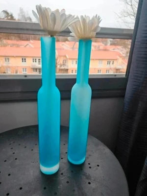 9 höga blå glasvaser - Säljer 9 stilrena, höga vaser i frostat blått glas med smal hals och cylinderformad kropp. Perfekta för enstaka blommor eller som dekorativa inredningsdetaljer. Ger ett modernt och fräscht intryck till rummet. Användes under en konfirmationsmottagning. Skickar med blomman i vaserna. Kan även hämtas i Partille