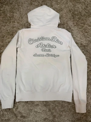 Vit hoodie från Christian Dior - Säljer en vit hoodie från Christian Dior med tryck 'Christian Dior Atelier Paris Avenue Montaigne' på både bröstet och ryggen. Hoodien har huva med dragsko och en stor magficka. Tillverkad i mjukt material och har ribbade muddar vid ärmar och nederkant.