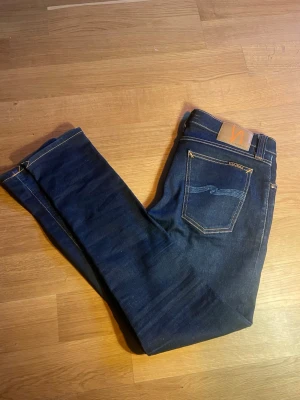 Mörkblå jeans från Nudie Jeans - Säljer ett par mörkblå jeans från Nudie Jeans med klassisk femficksmodell och kontrastsömmar i orange. Jeansen har raka ben och en snygg våglik logga på bakfickan. Perfekta för dig som gillar stilrena och tidlösa jeans.