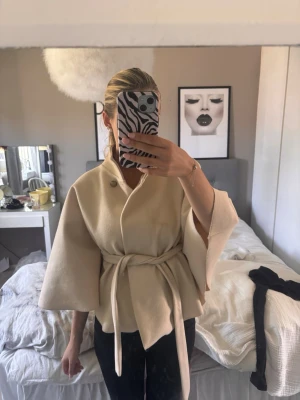 Beige kappa med knytband och vida ärmar - Stilren beige kappa med knytband i midjan och vida trekvartsärmar. Kappan har hög krage och stängs med en knapp upptill. Materialet ser ut att vara mjukt och tjockt, perfekt för kyligare dagar. Passar dig som gillar minimalistisk och trendig stil. Kappan har en fläck där framme som nästan knappt märks!! (har inte försökt att tvätta)