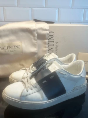 Valentino vita sneakers med svart detalj - Säljer ett par vita Valentino Garavani sneakers med bred svart läderdetalj över snörningen och ikoniska nitar på hälen. Skorna är köpta på merchsweden för ungefär 6000 tusen kronor för 1 år sedan, skorna är i fint skick över funderingar är det bara att höra av er🤗