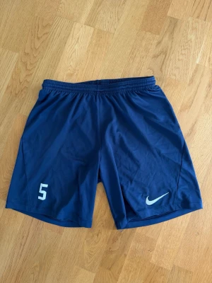 Blå Nike träningsshorts M - Säljer ett par marinblå träningsshorts från Nike med elastisk midja och siffran 5 tryckt på ena benet. Perfekta för fotboll eller annan träning. Lätta och bekväma med klassisk Nike-logga på benet.