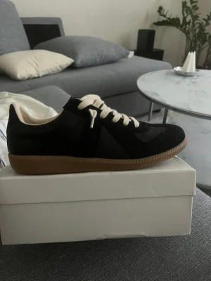 Svarta sneakers - Svarta sneakers med beige detaljer och gummisula. Skon har en klassisk låg silhuett, vita snören och ovandel i mocka och läder. Perfekt för dig som gillar stilrena och tidlösa sneakers med en lyxig touch.