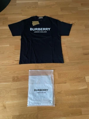 Svart Burberry t-shirt  - Säljer en svart t-shirt från Burberry med stor vit logga och texten 'London England' på bröstet. Klassisk rund hals och korta ärmar. Tillverkad i mjuk bomull och levereras med originalpåse och tagg. Denna t shirt och den vita jag även säljer kan köpas för 699 som ett paket pris. Tröjorna är i storlek S men passar mera M.