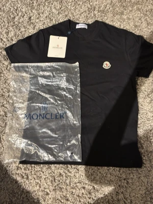 Svart Moncler t-shirt  - Snygg svart t-shirt från Moncler med klassisk rund hals och Moncler-logga på bröstet. Tillverkad i mjuk bomull för en skön känsla. Perfekt basplagg för dig som gillar stilrena och exklusiva detaljer.